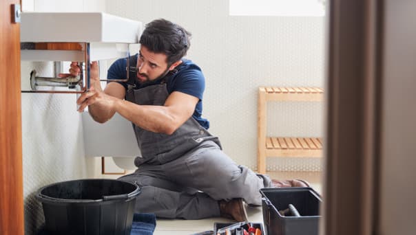 A plumber in overalls kneels on a bathroom floor, working on mechanical pipes under a sink with tools and a black bucket nearby.