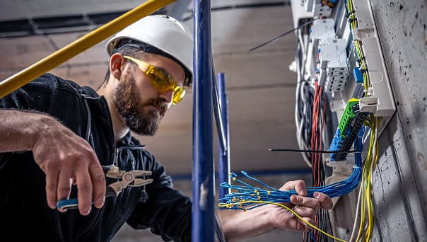Electrician wearing safety gear works with wire cutters and cables at an electrical panel in an industrial construction setting.