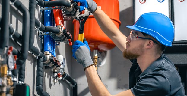 A worker wearing a blue hard hat and gloves uses a wrench to adjust mechanical plumbing pipes in an industrial construction setting.