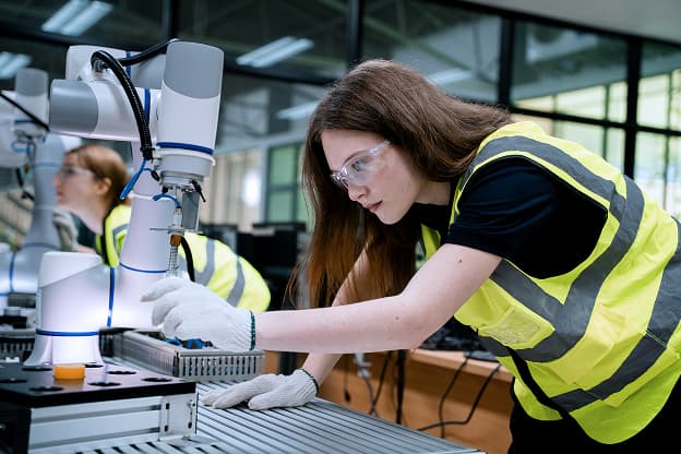 A woman wearing a high-visibility vest and safety glasses operates a robotic arm in an industrial MEP construction setting.