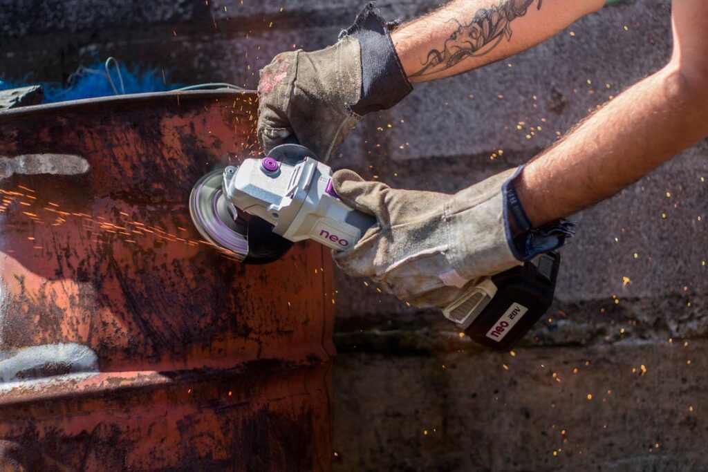 A person wearing gloves uses a handheld angle grinder to cut into a rusty metal barrel at a Colorado construction site, with sparks flying from the mechanical cutting area.