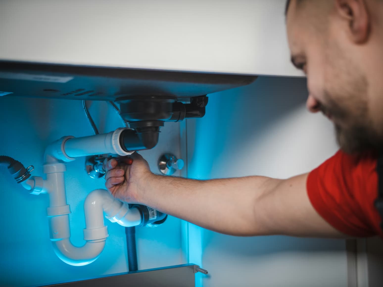 Person fixing white PVC pipes under a sink, adjusting plumbing connections with their hand, illuminated by blue light—a glimpse into Colorado plumbing at work.