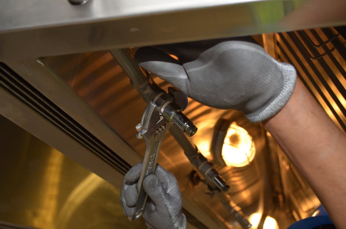 A person wearing gray gloves uses a wrench to tighten or loosen a pipe fitting inside a metallic industrial plumbing setting.