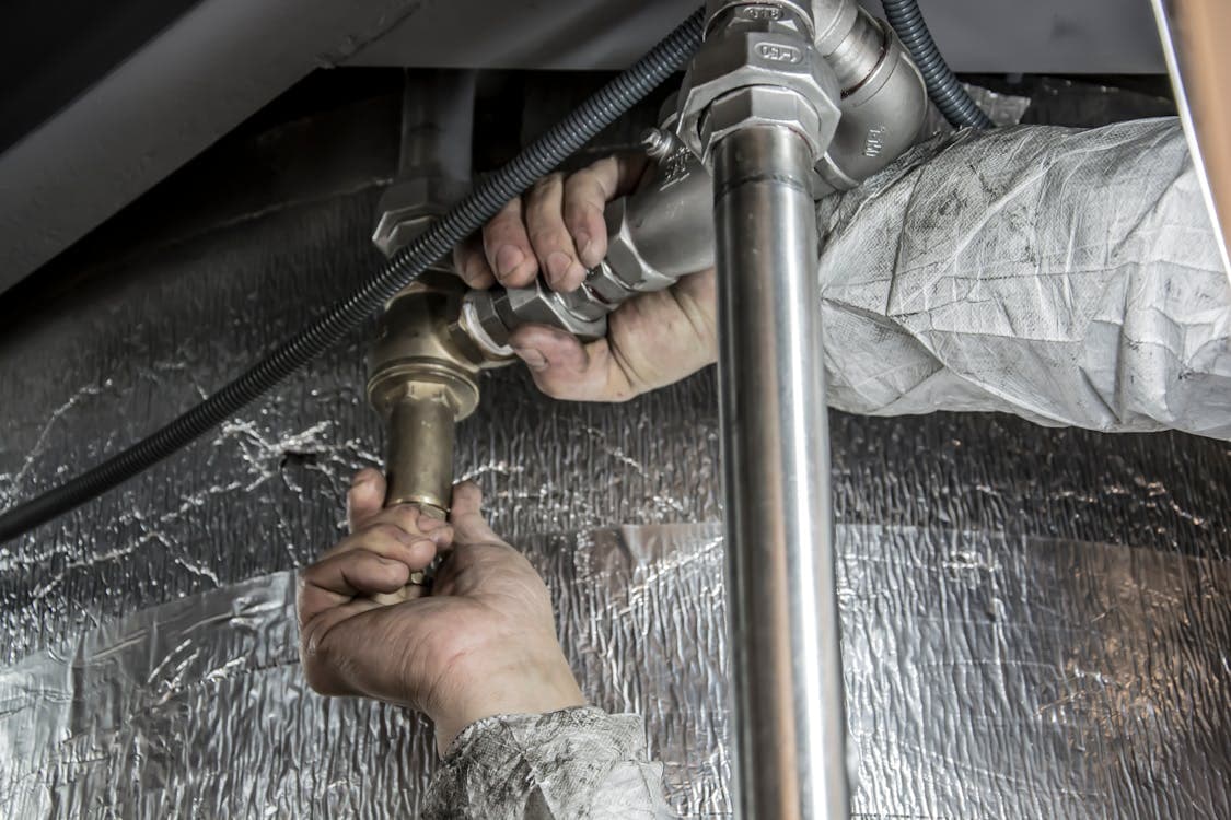 A person in protective clothing is repairing or installing metal plumbing pipes using both hands under a reflective insulated surface, showcasing skilled MEP work in construction.