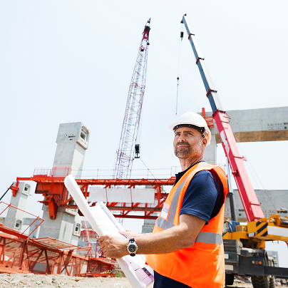 Man in safety gear holding blueprints at a construction site with cranes and concrete structures, overseeing mechanical, electrical, and plumbing installations.