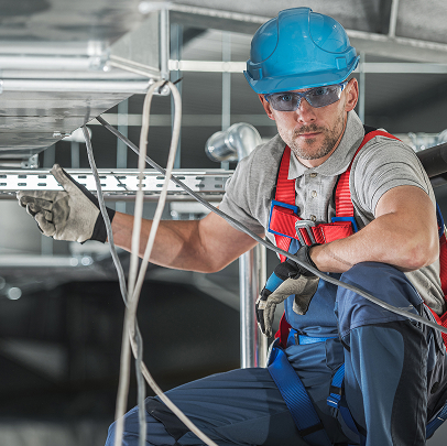 A construction worker in safety gear and helmet is inspecting or installing metal cables, ducts, and MEP components in an industrial setting.