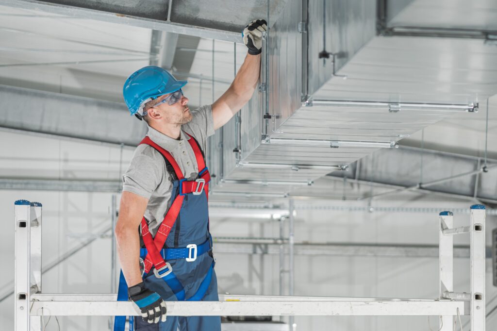 Worker wearing safety gear stands on a platform and inspects or installs a large metal duct system in an industrial building for a Colorado MEP project.
