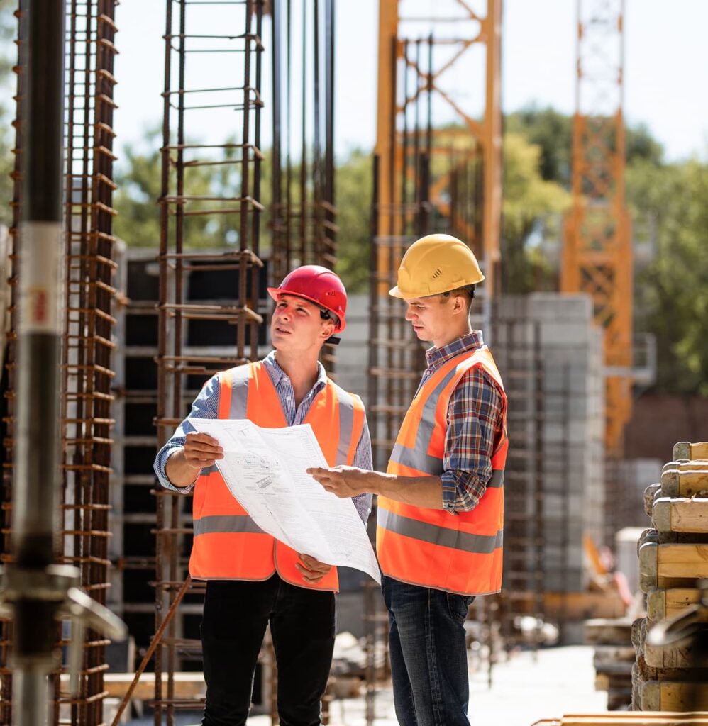 Two construction workers wearing safety vests and helmets examine mechanical blueprints at a Colorado building site with steel reinforcements and cranes in the background.