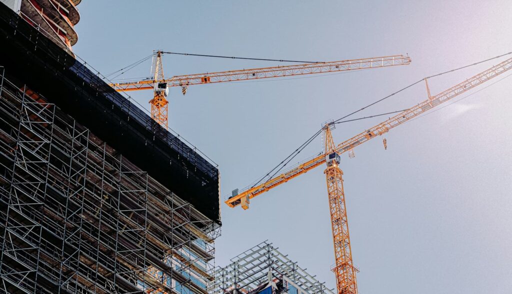 Two yellow construction cranes stand above scaffolding and a partially constructed building, where plumbing and electrical systems are being installed under a clear sky.