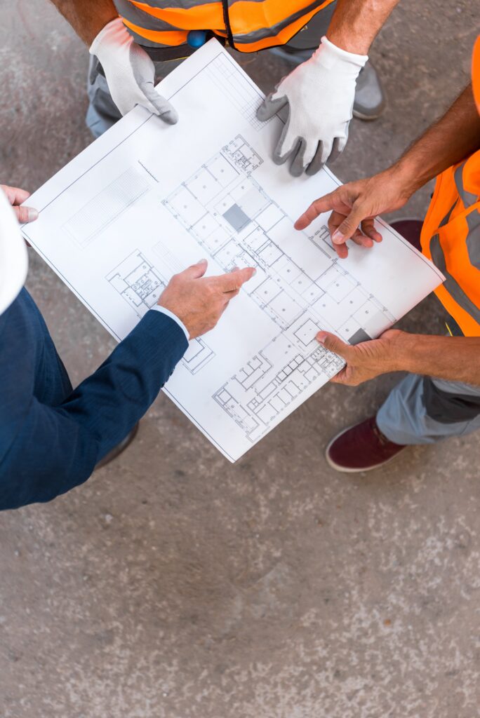 Three people, two in orange safety vests and one in a suit, review a construction floor plan on-site, pointing at different sections of the blueprint to discuss electrical and MEP details.