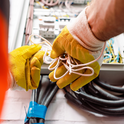 Two people wearing yellow work gloves handle electrical wires next to an open electrical panel, with various MEP components and cables visible.