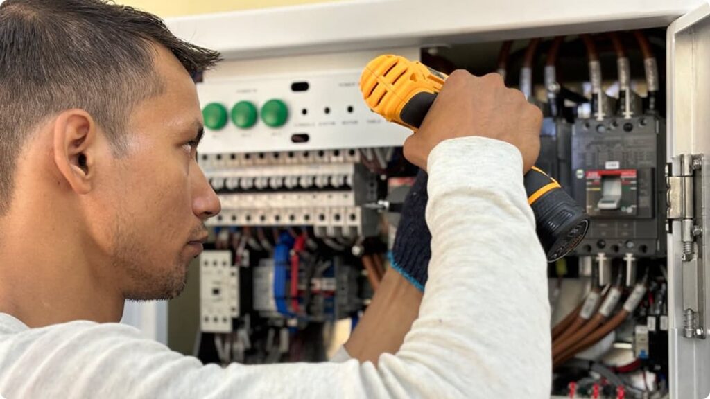 A person uses a yellow electric screwdriver to work on electrical wiring inside an open electrical control panel.