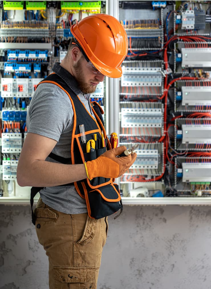 Electrician wearing a hard hat and safety vest checks a tool while standing in front of an open electrical panel with visible wiring, showcasing essential MEP work on a construction site.