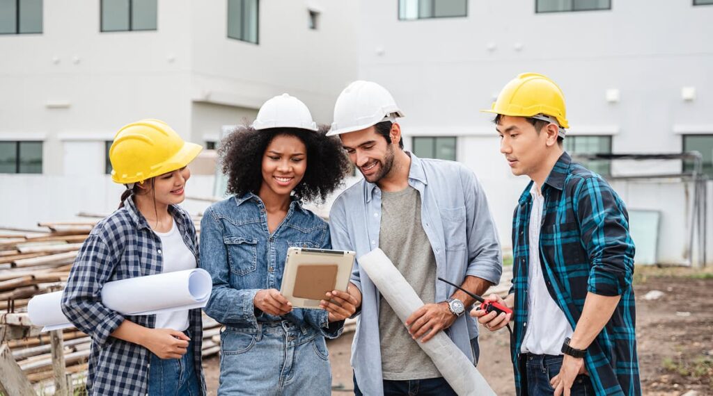 Four construction workers wearing safety helmets review electrical plans on a tablet at a Colorado building site, holding blueprints and tools, with a modern building in the background.