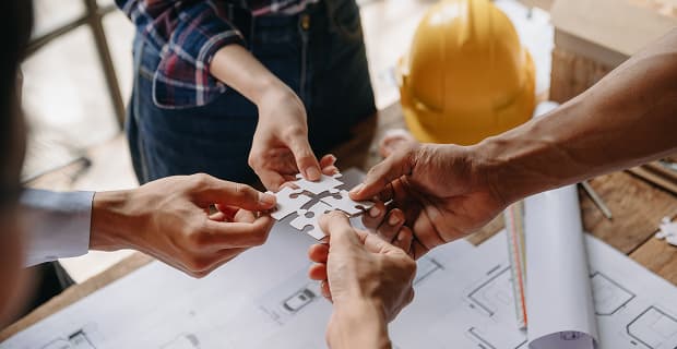 Four people hold puzzle pieces together over architectural blueprints in a Colorado construction setting, with a yellow hard hat, electrical plans, and rolled-up drawings visible on the table.