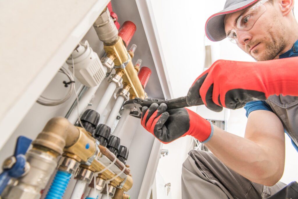 A plumber specializing in MEP services, wearing safety gear, uses a wrench to adjust pipes and valves inside an electrical control panel.