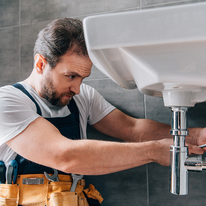 A plumber specializing in plumbing and MEP services is wearing a tool belt while repairing pipes under a white bathroom sink.