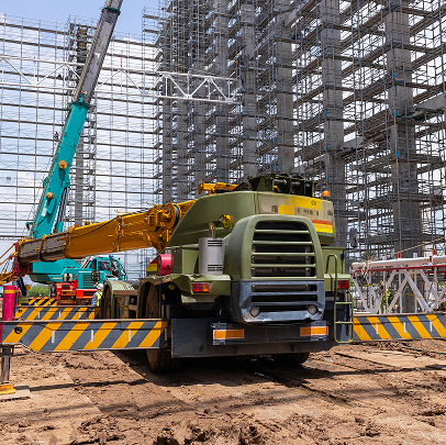 A construction site with a large green and yellow crane positioned on muddy ground, surrounded by metal scaffolding, building frameworks, and ongoing electrical work.