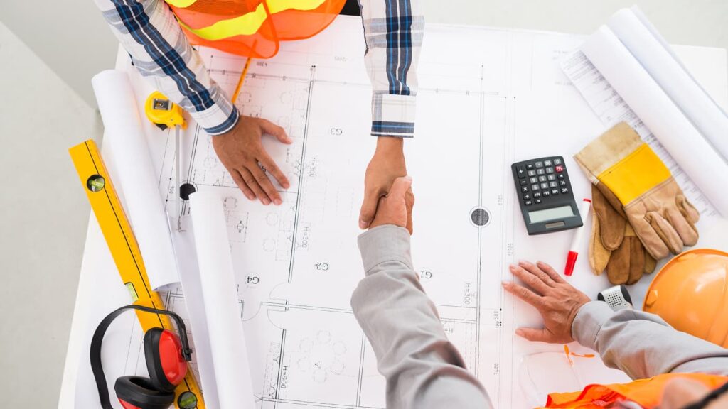 Two people shaking hands over architectural blueprints on a desk with construction tools, safety gear, a calculator, and electrical plans—signifying the start of a new construction project.
