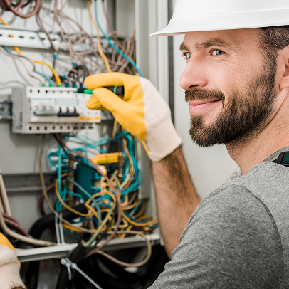 Electrician wearing gloves and a hard hat works on electrical wiring inside an MEP panel in Colorado, looking at the camera and smiling.