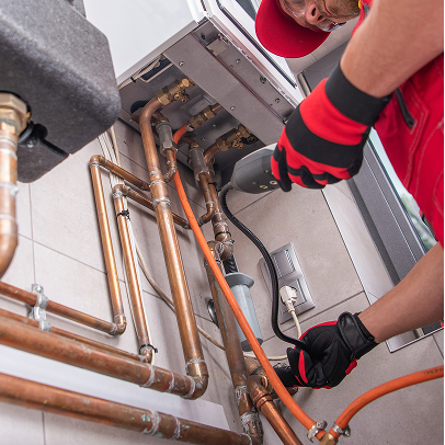 A technician in a red uniform inspects and services copper pipes and electrical equipment connected to a wall-mounted appliance, ensuring everything meets Colorado construction standards.