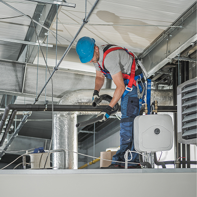 A worker wearing a safety harness and blue helmet is inspecting or repairing mechanical pipes near a ceiling in an industrial building.