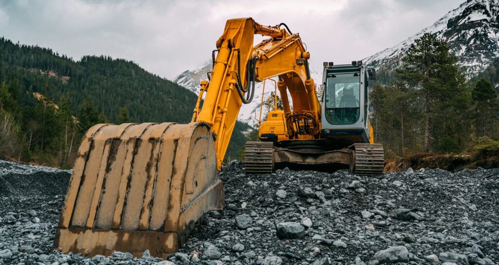 A large yellow excavator sits on rocky ground in a mountainous, forested Colorado landscape under a cloudy sky.