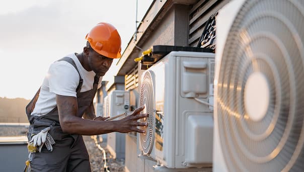 A technician in a hard hat and overalls inspects an outdoor air conditioning unit on a rooftop at a construction site in Colorado.