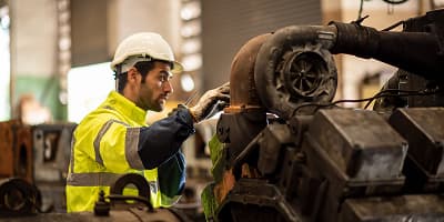 A worker in a high-visibility jacket and hard hat inspects or repairs MEP industrial machinery indoors, ensuring mechanical systems in Colorado facilities run smoothly.