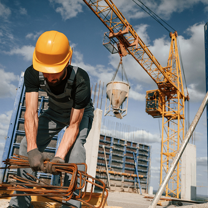 Construction worker in a yellow hard hat bends rebar at a Colorado building site, with mechanical equipment, a crane, and concrete bucket in the background under a blue sky.
