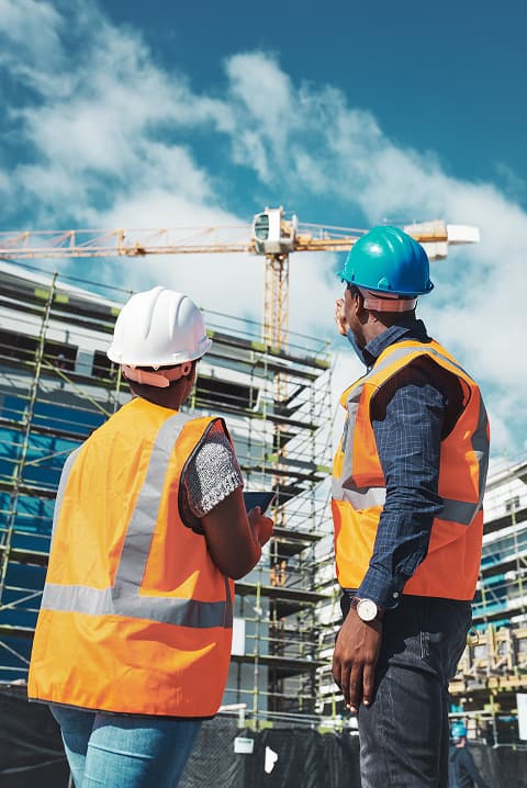 Two construction workers wearing safety vests and helmets observe a building site with scaffolding, a crane, and ongoing MEP and plumbing work in the background under a blue sky.
