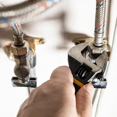 A hand uses a wrench to tighten a nut on a braided metal water supply hose connected to a valve during plumbing work.