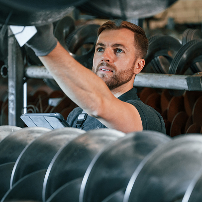 A man in work attire checks electrical inventory on shelves filled with metal pipes while holding a tablet.