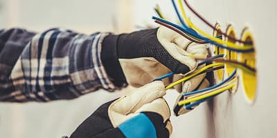 Person wearing gloves and a plaid shirt working with colored electrical wires in wall sockets, focusing on electrical tasks.