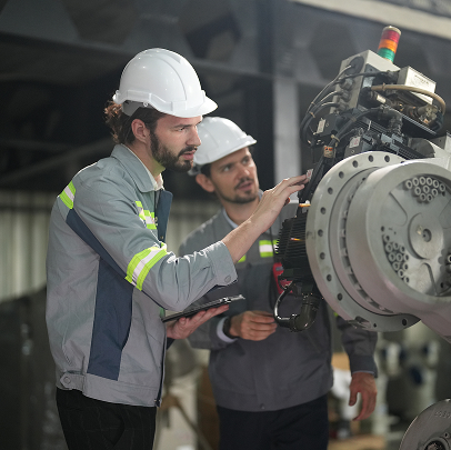 Two workers wearing hard hats and safety jackets inspect and operate a large industrial machine inside a construction or electrical factory setting.