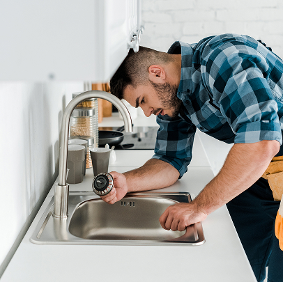 A man in a plaid shirt is inspecting a pipe under a kitchen sink, appearing to perform plumbing maintenance or mechanical repairs.