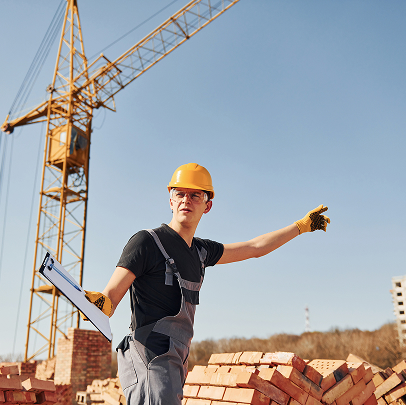 Construction worker wearing a yellow hard hat and gloves holds a clipboard and points, standing near bricks with a crane in the background under a clear sky, overseeing electrical or plumbing work on site.