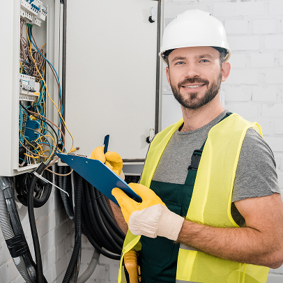 An electrician wearing a hard hat and safety vest inspects electrical wiring in a panel while holding a clipboard and pen at a construction site, ensuring all MEP systems meet safety standards.