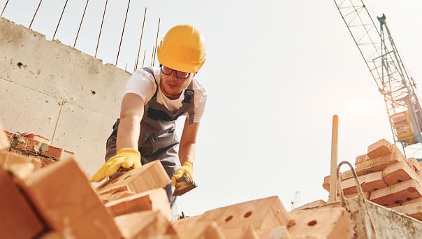Construction worker wearing a yellow hard hat and gloves is stacking bricks at a construction site under bright sunlight, preparing the area for upcoming electrical and MEP installations.