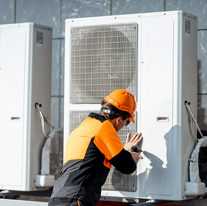Worker in safety gear repairs or inspects an outdoor air conditioning unit mounted on a building exterior, ensuring all electrical connections meet construction standards.